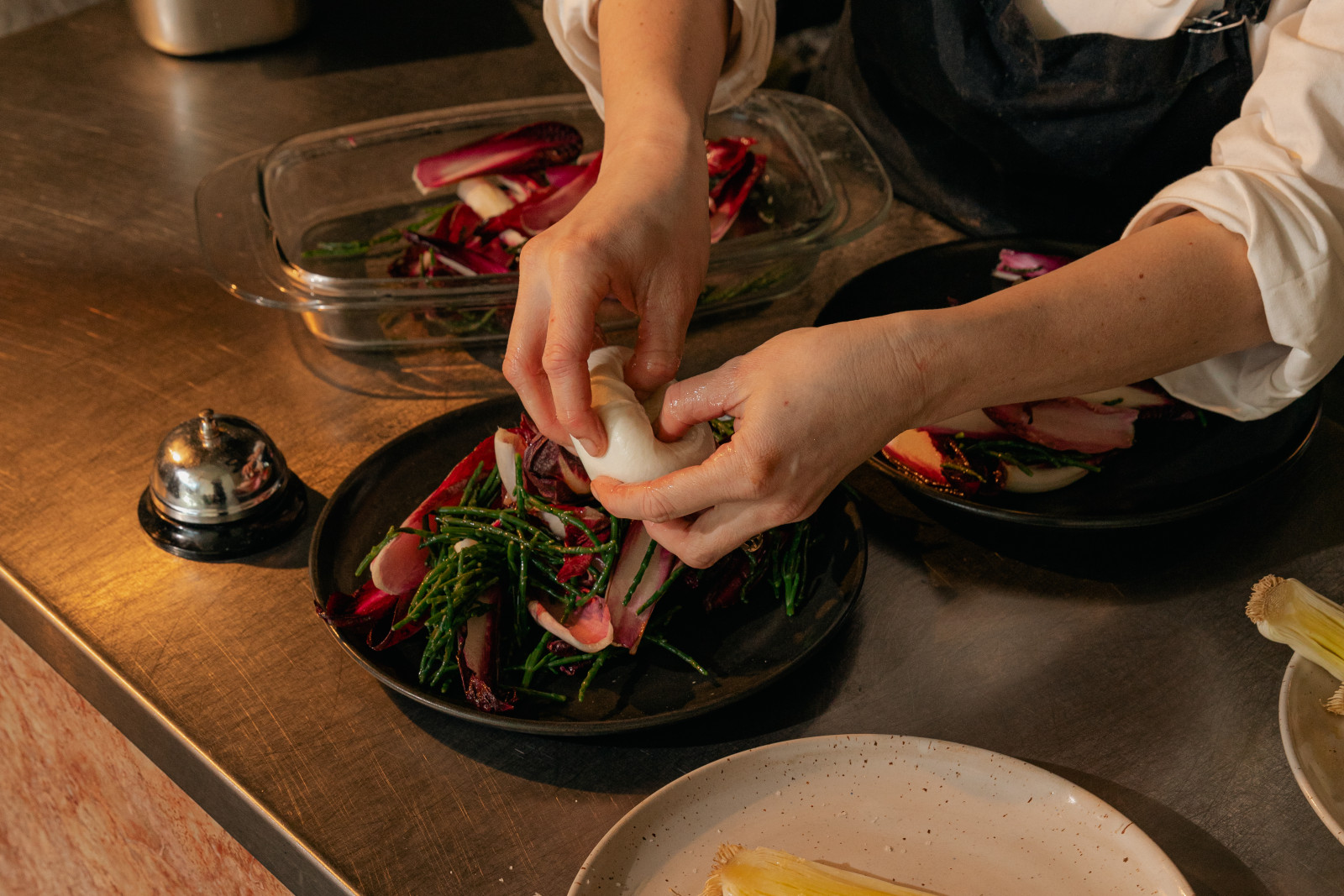 A cook plating burrata on a salad at the pass, a service bell beside the plate